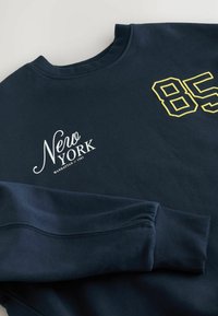 Sweatshirt bleu marine avec le texte "Nero York Manhattan // 1985" en blanc et le numéro "85" en contour jaune sur la poitrine, avec une manche repliée visible.