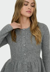 Siv kardigan z zrnato pletenjem, okroglim ovratnikom, dolgimi rokavi in sprednjim zapiranjem na gumb. Ima razširjen spodnji del.