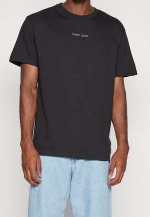 Mies, jolla on musta lyhythihainen T-paita, jossa on "TOMMY JEANS"-teksti keskellä rinnassa, ja vaaleansiniset farkut.