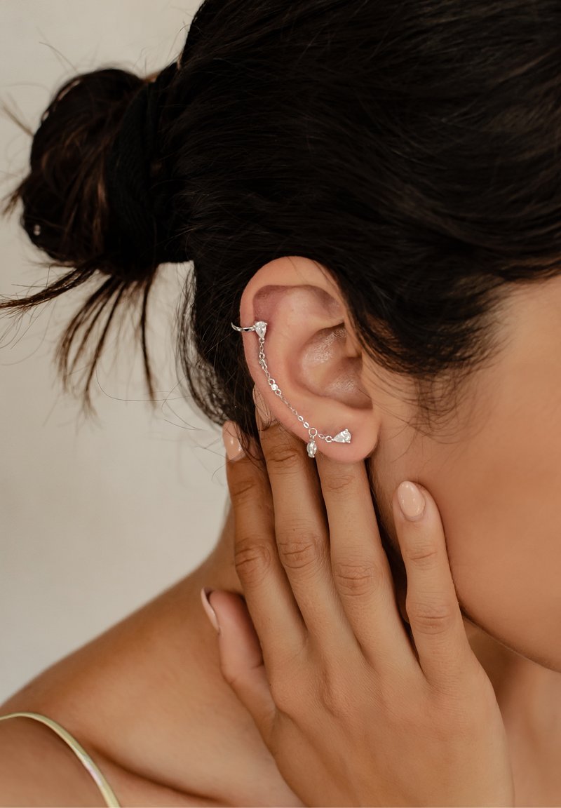 Femme aux cheveux foncés en chignon porte une délicate manchette d'oreille en argent reliée par une chaîne à une puce d'oreille, touchant son oreille avec une main manucurée.