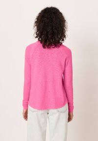 Osebek s kodrastimi lasmi, ki nosi dolgo srajco svetlo rožnate barve in svetlo modre kavbojke, stoji z obrazom obrnjenim proti kameri.