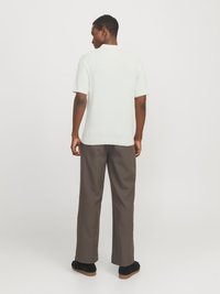 Bărbat cu fața spre spate, purtând o cămașă albă cu mânecă scurtă, pantaloni gri cu picioare largi și pantofi negri, pe un fundal simplu.