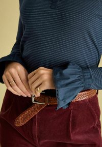 Marineblau gestreiftes Langarmhemd mit Rüschensaum, kombiniert mit burgunderfarbener Cordhose und einem gewobenen braunen Leder-Gürtel mit einer silbernen Schnalle.