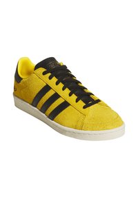 Gelber Wildleder-Sneaker mit schwarzen Lederakzenten und Drei-Streifen-Design. Strukturierte Oberseite, schwarze Schnürsenkel und weiße Gummisohlen-Details.