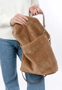 Braune Kunstfelltasche mit glatter Textur, einem braunen Ledertrageriemen und einem geräumigen Innenraum, gehalten von einer Person in hellen Jeans.