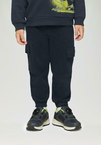 Marine cargobroek met elastische boorden en twee zijzakken, gedragen met zwarte sneakers met groene accenten en een bevestigingssysteem.