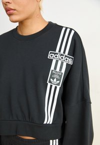 Melna īsa sporta jaka ar baltu Adidas logotipa uzšuvumu un trim baltām svītrām uz piedurknēm. Mīksts audums ar ribu apakšmalu un piedurkņu malām.
