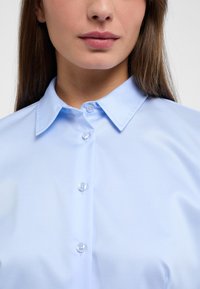 Light blue collared shirt made of smooth fabric, featuring a button placket with clear buttons and a clean, fitted design.