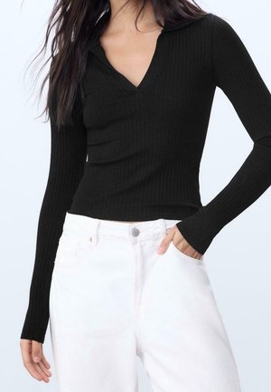 Femme portant un haut noir côtelé moulant à manches longues avec un petit col en V, associé à un pantalon blanc taille haute, une main dans la poche.