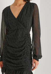 Vestido negro con lunares blancos, mangas largas transparentes, escote en V y textura fruncida. El detalle del dobladillo con volantes añade dimensión al diseño.