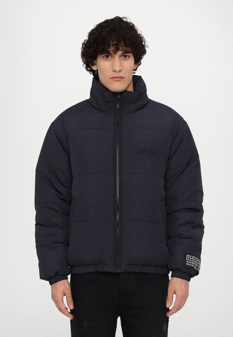 Veste matelassée navy avec col montant, fermeture éclair, motif matelassé et manchons en tricot dotés d'un accent clouté au poignet.
