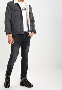 Chaqueta de mezclilla gris con cuello de borrego blanco, camiseta blanca, pantalones vaqueros negros y botas de cordones marrón oscuro. Corte ajustado con bolsillos frontales.