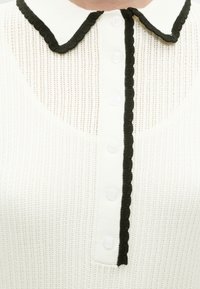 Κοντινή λήψη λευκού πλεκτού τοπ με μαύρο κυματιστό περίγραμμα στη γιακά Peter Pan και στη λωρίδα με κουμπιά.