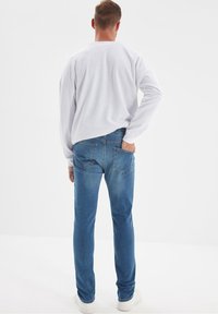 Jean bleu à coupe régulière avec des accents de surpiqûres, associé à une chemise blanche à manches longues et des baskets blanches. Tenue simple et décontractée.