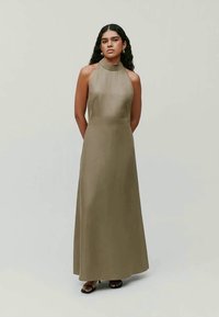 Femeie cu păr lung și închis la culoare, purtând o rochie fără mâneci, verde măsliniu, cu guler înalt și lungime până la podea, și sandale negre cu vârf deschis, stând cu mâinile în spate.