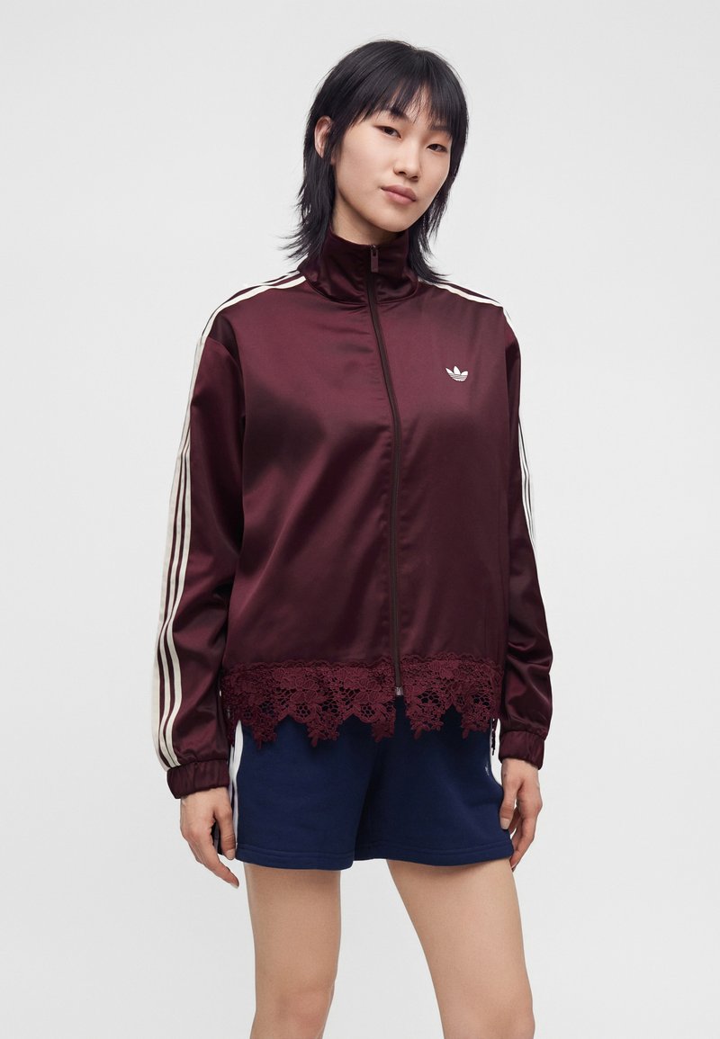 Jeune femme portant une veste zippée bordeaux avec des rayures blanches sur les manches et un bord en dentelle, associée à un short de sport bleu marine.