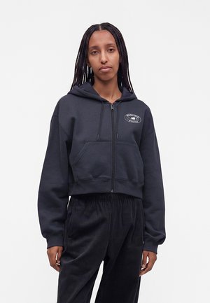 Woman with long braids wearing a dark zip-up hoodie with front pockets and black corduroy pants standing against a plain background.