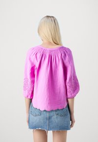 Blusa rosa con scollo arricciato e maniche a palloncino, caratterizzata da accenti floreali ricamati. Orlo a onda e tessuto leggero.