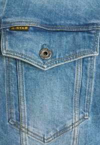 Detalj av en jeansjacka i ljusblått, med etikett i gult "G-STAR", silverfärgad knapp, kontrasterande sömmar och en design med spetsig ficklock.