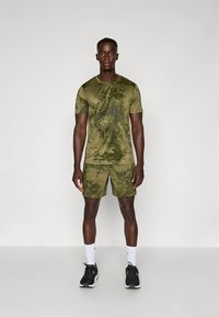 Oliiviroheline spordisärk ja lühikesed püksid kamuflaažimustriga; kerge ja keha järgnev disain; kombineeritud mustade tossudega ja valgete sokkidega.