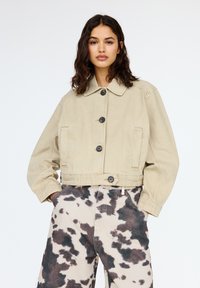 Giacca corta beige con colletto, chiusura con bottoni e due tasche laterali; abbinata a pantaloni beige chiaro con una stampa di mucca marrone scuro.