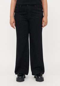Person wearing black wide-leg pants with subtle leopard print and black chunky-soled shoes, standing against a white background.