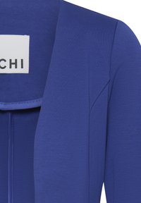 Primo piano di un blazer blu vivace che mostra il rever, la cucitura della spalla e un'etichetta bianca parziale con testo nero "CHI".