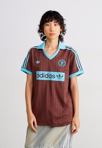Osoba s krátkymi kučerami má na sebe hnedé športové tričko Adidas so svetlomodrým golierom, pruhmi a logom, doplnené svetlošedou sukňou.