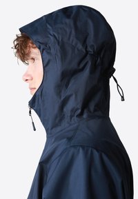 Chaqueta cortavientos de color marino con capucha ajustada, textura suave, cremallera frontal y cordón ajustable. Presenta un diseño minimalista y costuras.