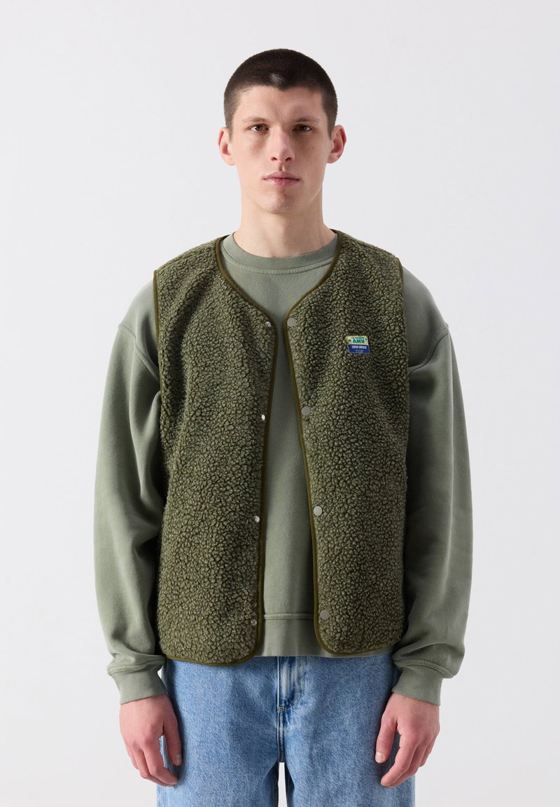 Mlad moški stoji obrnjen proti naprej, oblečen v olivno zelen flis brezrokavnik čez ujemajočo se trenirko in svetlo modre kavbojke na preprostem belem ozadju.