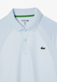 Ljusblå polo-shirt med en ribbad krage, tvåknappsfäste och mesh-sidopaneler. Har en liten grön krokodil-logotyp. Slim fit design.