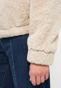 Μπεζ πουλόβερ από αφράτο ύφασμα με υφή, με ριμπ μανσέτες και στρογγυλεμένη κάτω ραφή, συνδυασμένο με σκουρόχρωμο τζιν.