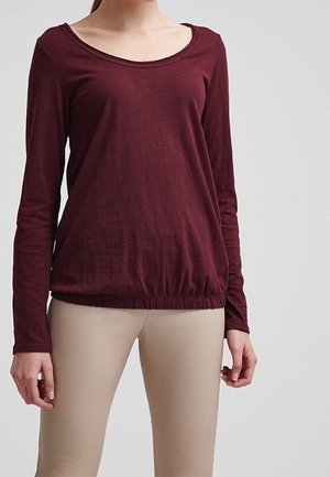 Kvinde iført en langærmet, vinrød top med rund hals og elastisk kant samt beige, stramme bukser foran en ensfarvet lys baggrund.