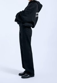 Svart oversized hoodie med vitt logotyp, kombinerad med svarta högmidjade byxor och svarta loafers. Slätt, mjukt tyg och avslappnad passform.