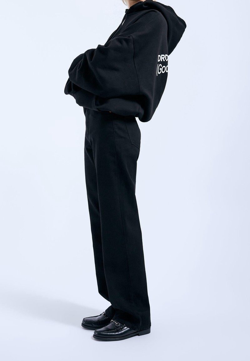 Svart oversized hoodie med vitt logotyp, kombinerad med svarta högmidjade byxor och svarta loafers. Slätt, mjukt tyg och avslappnad passform.