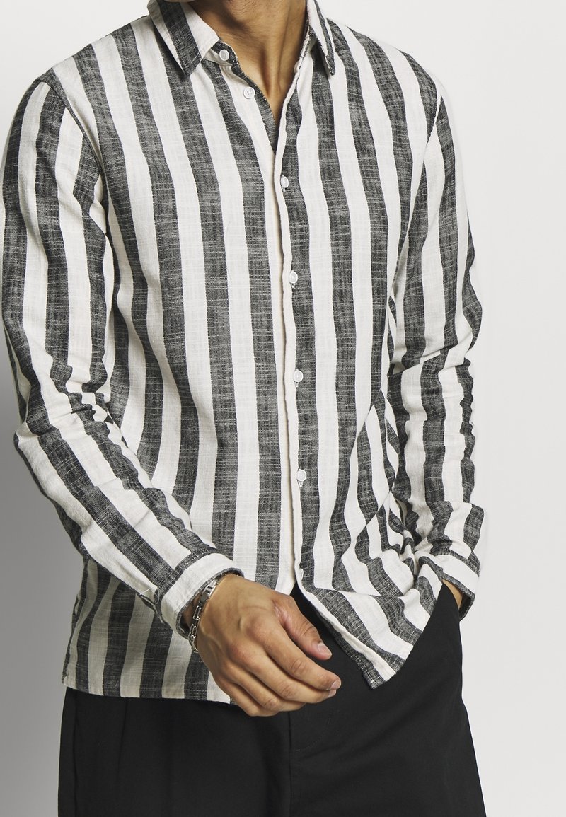 Homme portant une chemise blanche à manches longues avec des rayures noires verticales, un pantalon noir, un bracelet, et une main dans la poche, sur fond clair.