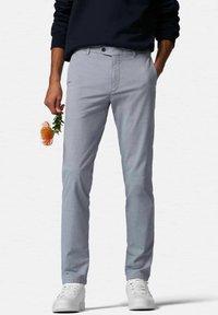 Hellgraue Chinos mit strukturierter Optik, schmaler Schnitt, Knopfverschluss und Fronttaschen, kombiniert mit weißen Sneakers. In einer Hand hält die Person eine Blume.