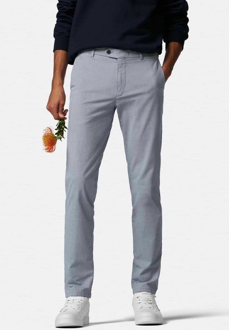 Hellgraue Chinos mit strukturierter Optik, schmaler Schnitt, Knopfverschluss und Fronttaschen, kombiniert mit weißen Sneakers. In einer Hand hält die Person eine Blume.