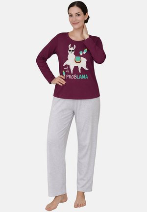 Woman wearing purple long-sleeve top with llama graphic and "not my PROBLAMA" text and light gray pajama pants, standing barefoot.