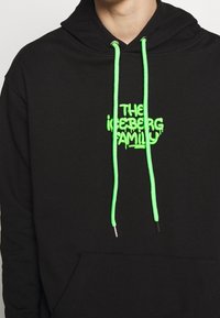 Svart huvtröja med en framficka, med neongröna snoddar och broderad text "THE ICEBERG FAMILY" i en droppande font.