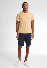 Camiseta de algodón beige con mangas cortas y un bolsillo en el pecho, combinada con pantalones cortos azul marino. El modelo lleva zapatillas blancas y está de pie sobre un fondo liso.