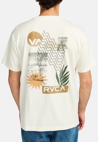 Cream-colored T-shirt featuring multicolored graphic print with abstract designs, a palm leaf, and brand logos in gold and green accents.