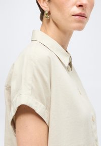 Femme portant une chemise beige à manches courtes avec col et une boucle d'oreille créole dorée texturée, vue de l'épaule vers le haut sur fond blanc.