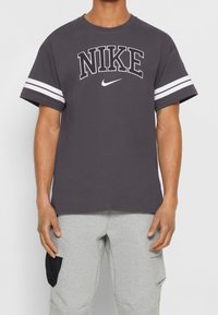 Šedé bavlněné tričko s bílým logem "NIKE" a pruhy na rukávech, kulatý výstřih, volný střih, hladká textura, krátké rukávy.