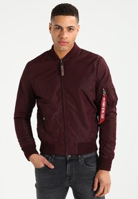 Jachetă bomber maro cu manșete și tiv ribbed, fermoar în față, buzunare laterale și un buzunar utilitar pe mânecă cu un etichetă roșie.