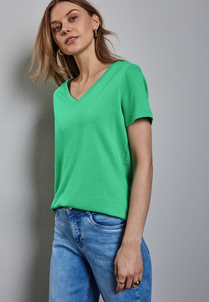 Zelené tričko s výstřihem do V z měkké bavlny s krátkými rukávy, spárované se světlými modrými džíny, nošené modelem na neutrálním pozadí.