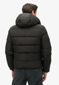 Sort pufferjakke med høj krave og en syet design. Har et logo på ryggen og tilbyder en komfortabel og isoleret pasform.