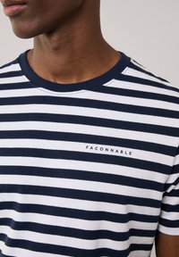 T-shirta z dekoltem okrągłym w biało-granatowe paski, wykonana z bawełny, z małym logo na piersi. Gładka tekstura, luźny krój.