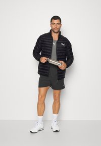 Sort pufferjakke med vandret syninger, grå atletisk tanktop og sorte shorts. Holder en sølvfarvet vandflaske, iført hvide sneakers.