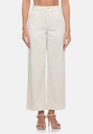 Donna che indossa pantaloni bianchi a vita alta e gamba larga abbinati a sandali beige con tacco aperto e cinturini alla caviglia.